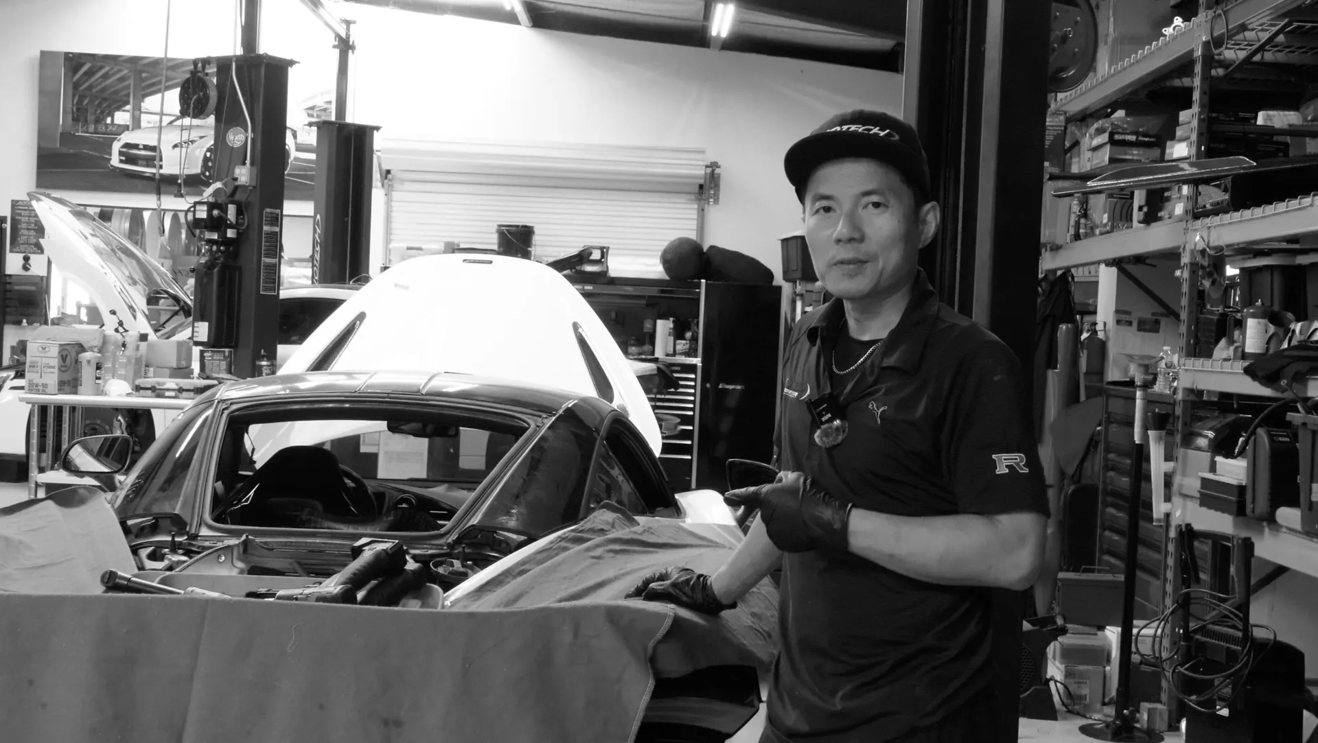 Mechanic in a Jotech Motorsports shop working on a car with the trunk open, surrounded by tools and equipment.